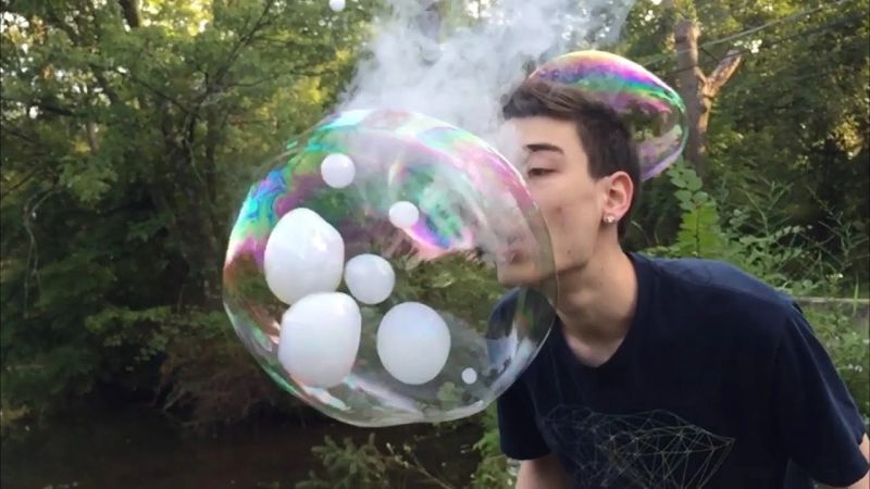 A young man blowing a large bubble filled with smoke and smaller bubbles inside it.